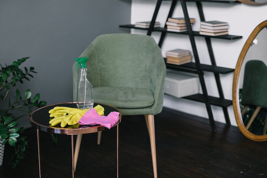 A person wearing a light green cleaning glove is reaching towards a wooden bookshelf with a dark finish, containing a row of green hardcover books. The individual is holding a white cloth or cleaning