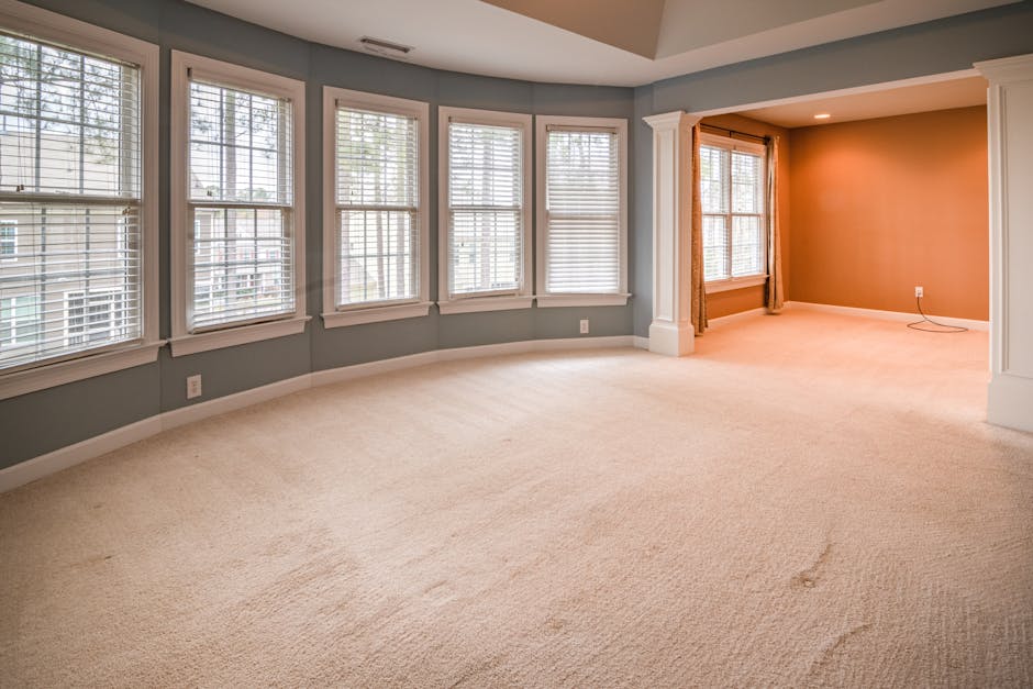 A person using a vacuum cleaner with a yellow body and black hose attachment to perform surface cleaning on a patterned, cream-colored carpet in a residential setting. The individual is dressed in cas